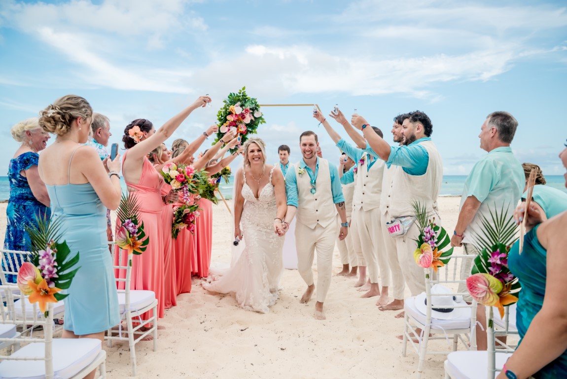 Destination wedding couple walking down the beach aisle with guests celebrating